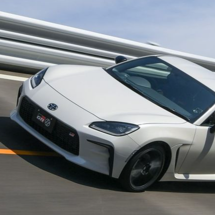 A white Toyota GR86 sports coupe speeding along a highway, captured from a low front-three-quarter angle. The car features aggressive styling with a black honeycomb grille, sharp LED headlights, and a prominent “GR” badge on the front bumper. It has black alloy wheels and aerodynamic side skirts. The background shows motion blur of the road and a metal guardrail under a bright blue sky, emphasizing speed and performance.
